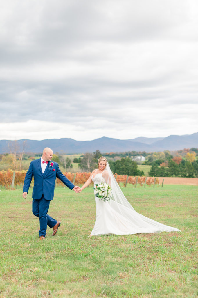 Groom in blue tux with red bowtie leading bride with blue and white bouquet across CrossKeys Vineyard taken by Ashley Eagleson Photography.