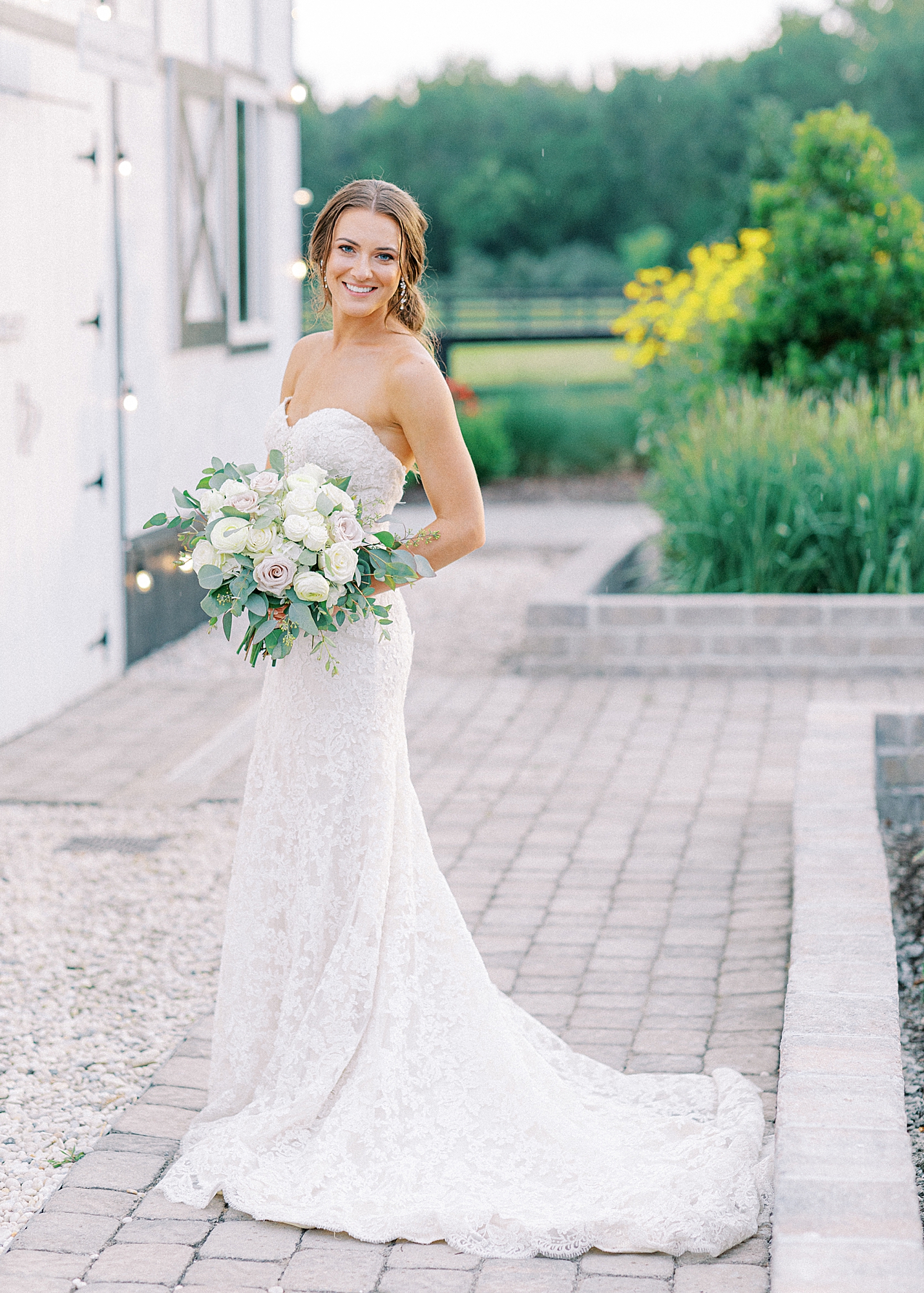 Bride wearing lace strapless wedding dress smiling into the camera holding white and light blush bouquet.