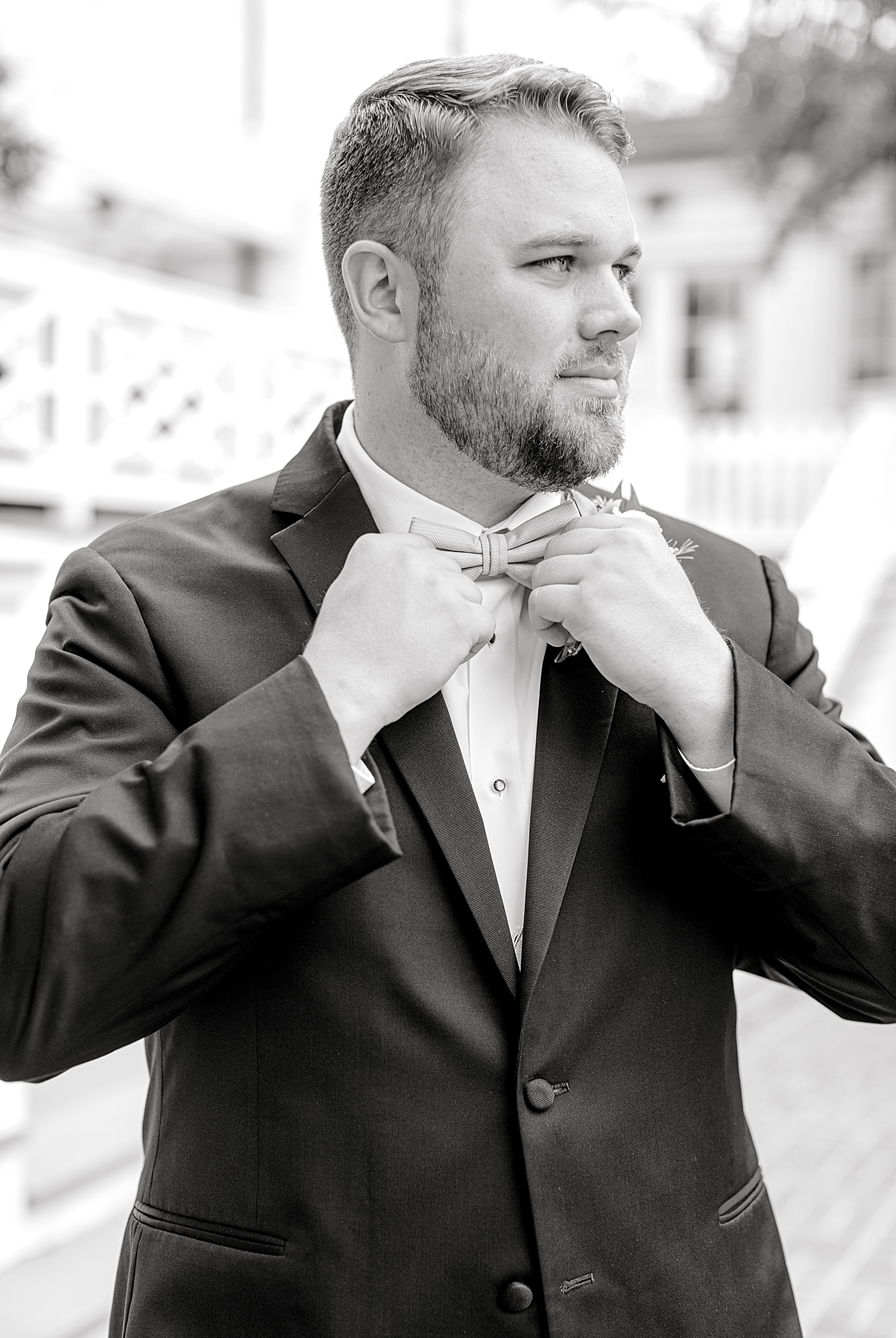 Black and white photo of groom fixing bowtie at estate.