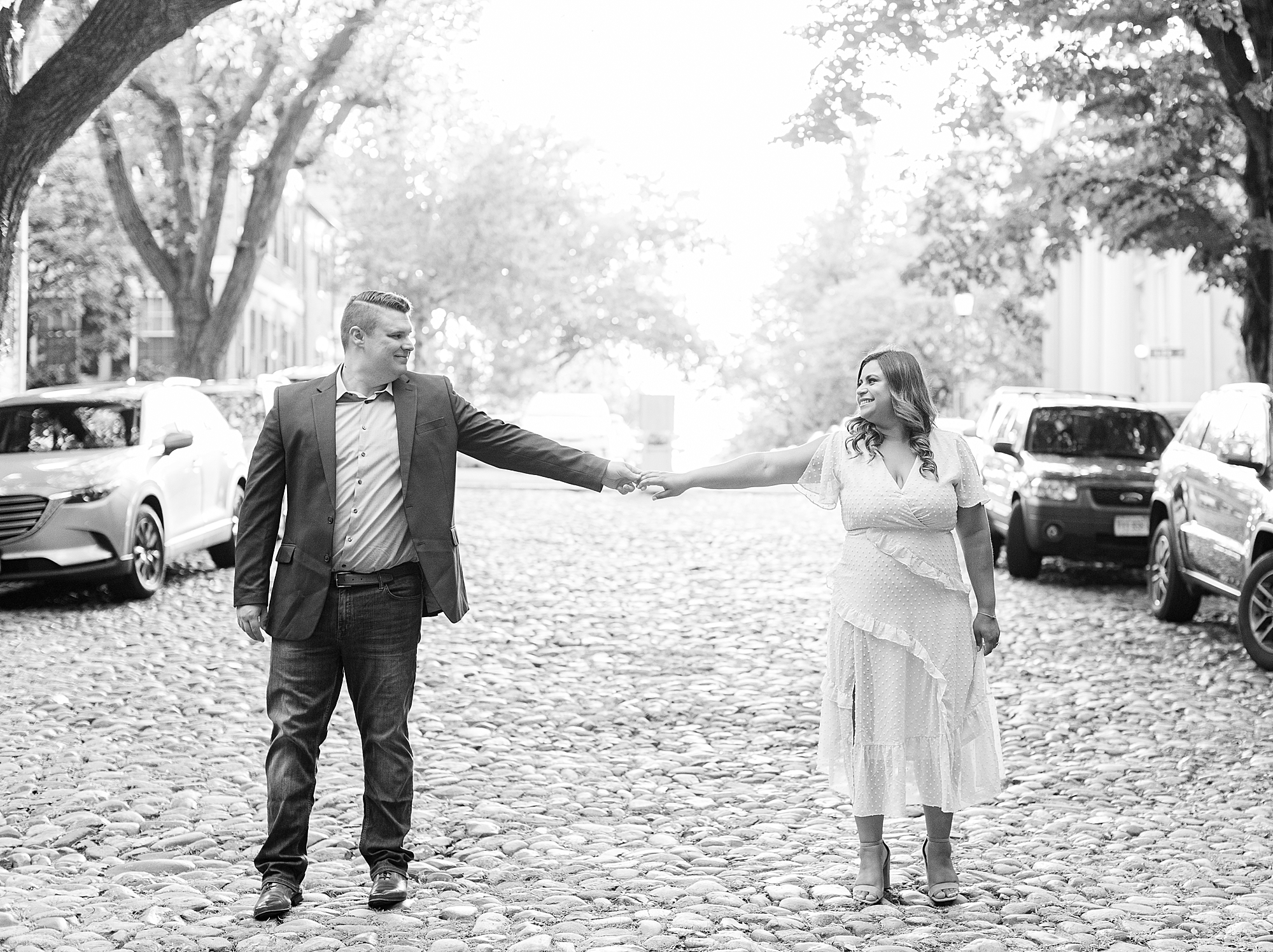 Cobblestone streets of Old Town Alexandria. Couple stretches out to hold hands during engagement session.