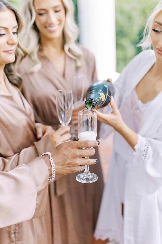 Bride pouring champagne in brown getting ready robe.