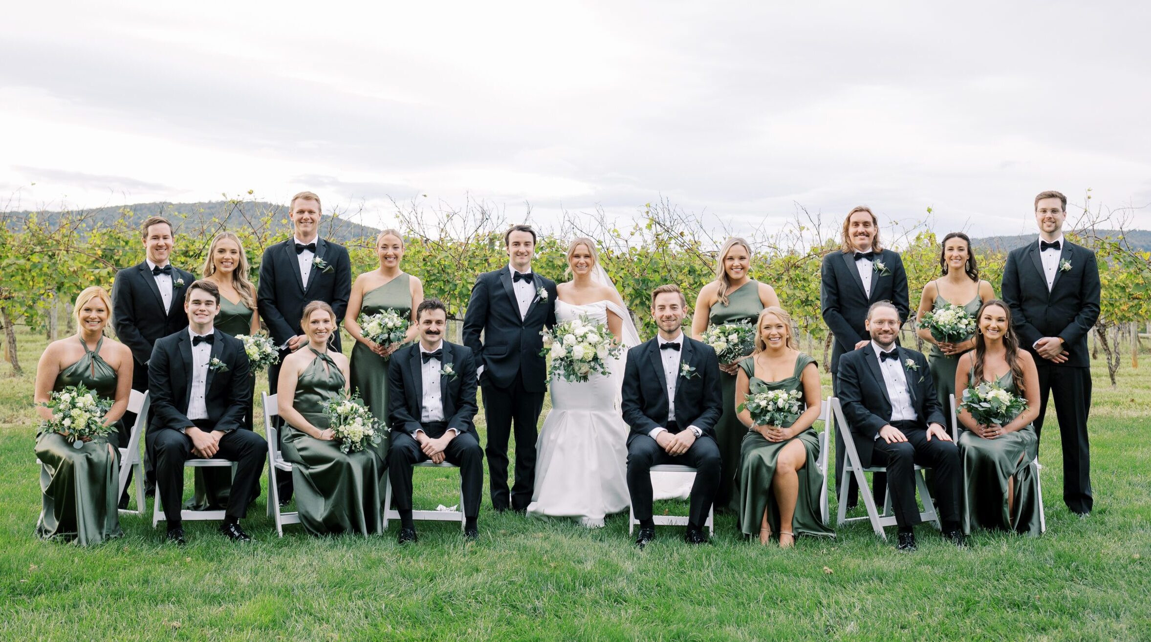 Group wedding party photo with bridesmaids sitting down in green satin dresses at Keswick Vineyards.